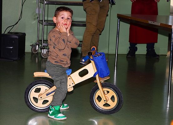 Ein junger Junge sitzt auf einem Holzfahrrad mit Stützrädern und hält sich die Hand vor den Mund. Ein Mensch in Hosen steht hinter ihm. Im Hintergrund befindet sich ein Tisch, ein Lautsprecher und ein Regal.