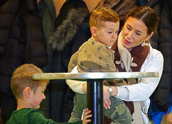 Eine Frau hält einen jungen Jungen in ihren Armen, während ein anderer Junge sie von unter einem Tisch aus beobachtet. Im Hintergrund hängen Winterjacken.