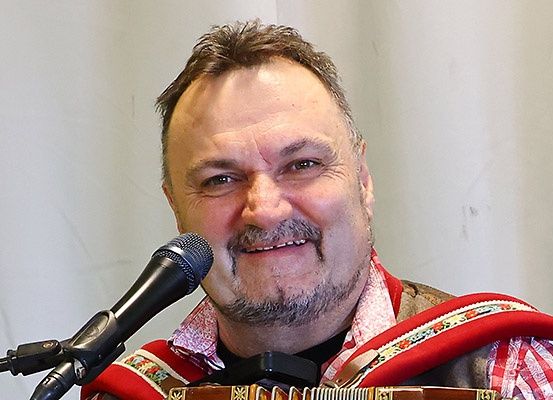 Ein Mann mit Bart lächelt und steht vor einem Mikrofon. Er trägt eine rot-weiße Weste und hat ein schwarzes Akkordeon über der Schulter.
