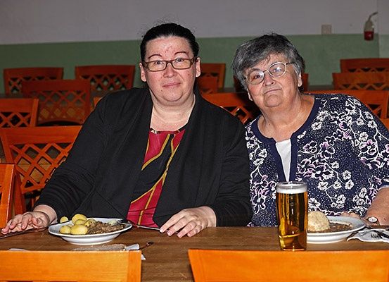 Zwei Frauen sitzen an einem Holztisch in einem Restaurant, lächeln und posieren für ein Foto. Eine hat eine Schüssel mit Essen und die andere ein Glas Bier.