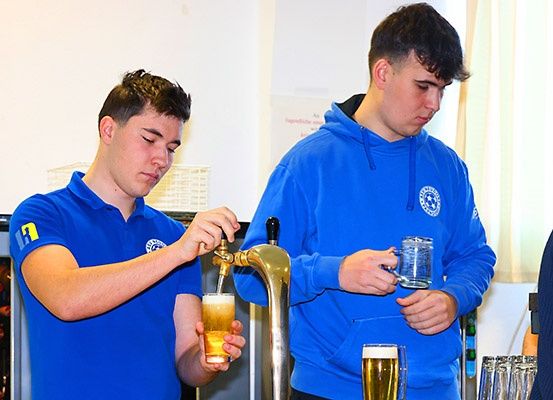 Zwei junge Männer in blauen Uniformen schenken Bier aus einem Zapfhahn ein. Einer hält ein Glas und der andere hat ein Becher. Eine weiße Wand ist hinter ihnen.