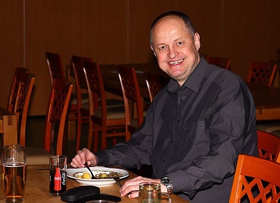 Ein Mann in einem schwarzen Hemd sitzt an einem Tisch, lächelt und hält eine Gabel. Er isst Essen aus einer Schüssel, mit einem Glas Bier und einer Coca-Cola-Dose auf dem Tisch. Im Hintergrund befinden sich Stühle und ein weiterer Tisch.