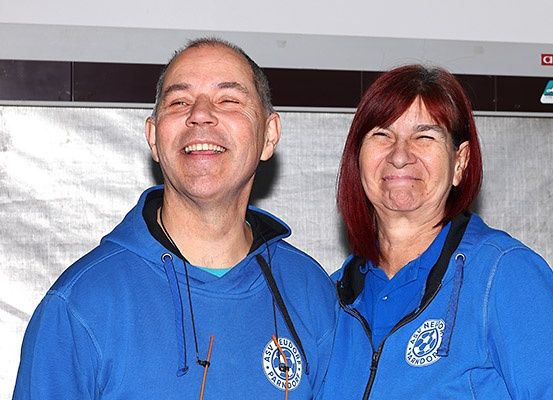 Ein lächelnder Mann und eine Frau stehen eng beieinander und tragen beide blaue Kapuzenpullover mit einem Logo auf der Vorderseite. Sie scheinen für ein Foto zu posieren.