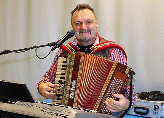 Ein Mann steht mit einem Akkordeon in den Händen an einem Mikrofon und trägt ein rotes kariertes Hemd, während ein Keyboard im Vordergrund steht.