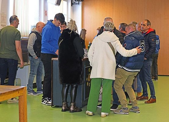 Eine Gruppe von Menschen steht in einer Schlange, wahrscheinlich in einem Wartebereich. Einige schauen nach unten, während andere in ein Gespräch vertieft sind. Der Raum hat Holzwände und einen Holztisch mit einem Glas.