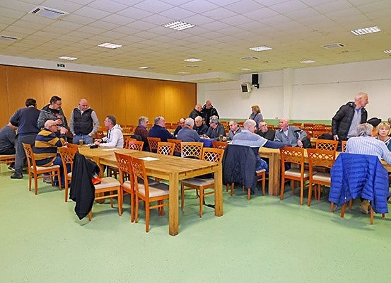 Eine Gruppe von Menschen versammelt sich in einem Raum mit Holztischen und Stühlen. Einige stehen und andere sitzen, alle führen Gespräche. Der Raum hat Holzwände, eine weiße Decke und einen grünen Boden. An der Wand ist ein Lautsprecher montiert.