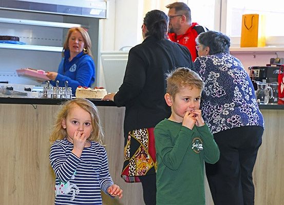 Eine Gruppe von Menschen steht an einem Tresen, wo eine Frau Kuchen serviert. Zwei Kinder schauen auf den Kuchen, ein Mädchen mit den Händen im Mund und ein Junge mit den Händen am Kinn. Hinter ihnen schaut eine Frau auf den Kuchen.