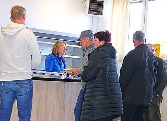 Mehrere Personen versammeln sich in einem Raum, möglicherweise einer Cafeteria, wo eine Frau Essen serviert. Ein Mann in einer grauen Mütze und einem Jackett unterhält sich mit einem anderen Mann. Dahinter spricht eine Frau in einer blauen Jacke mit einem Mann in einer weißen Kapuzenjacke. Eine Klimaanlage ist an der Wand montiert.
