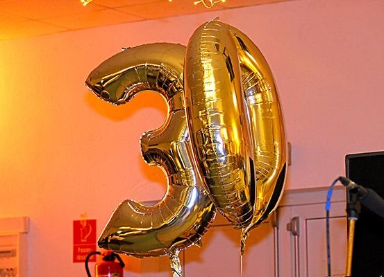 Zwei goldene Folienballons in Form der Zahl 30, an einer rosa Wand platziert. Links ist ein roter Feuerlöscher zu sehen, rechts steht ein Mikrofon.