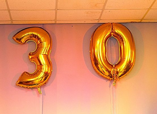 Zwei goldene Folienballons, einer mit der Nummer 3 und der andere mit der Nummer 0, hängen von der Decke an einer rosa Wand.