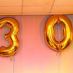 Zwei goldene Folienballons, einer mit der Nummer 3 und der andere mit der Nummer 0, hängen von der Decke an einer rosa Wand.