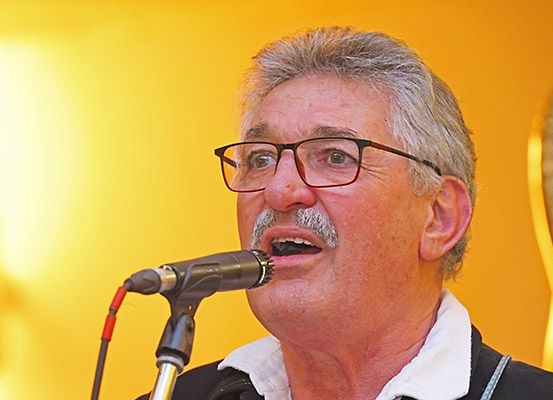 Ein älterer Mann mit grauem Haar und einem Schnurrbart singt mit ernster Miene in ein Mikrofon. Er trägt eine Brille und einen weißen Kragen. Hinter ihm befindet sich eine gelbe Wand und ein goldenes Objekt.
