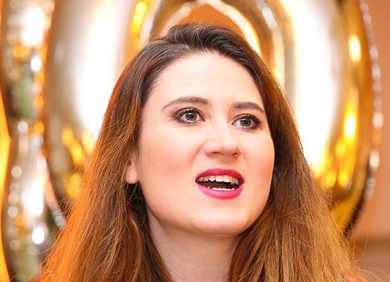 Eine Frau mit braunen Haaren und rosa Lippenstift lächelt mit offenem Mund. Sie steht vor einem goldenen Luftballon.