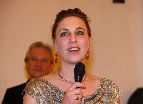 Eine Frau in einem goldenen Kleid hält ein Mikrofon und lächelt in die Kamera, mit einem Mann hinter ihr.