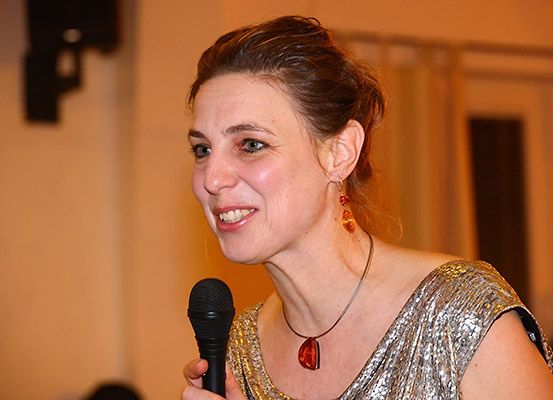 Eine lächelnde Frau mit braunen Haaren, die ein goldfarbenes Paillettenkleid und Ohrringe trägt, spricht in ein schwarzes Mikrofon. Der Hintergrund ist unscharf.