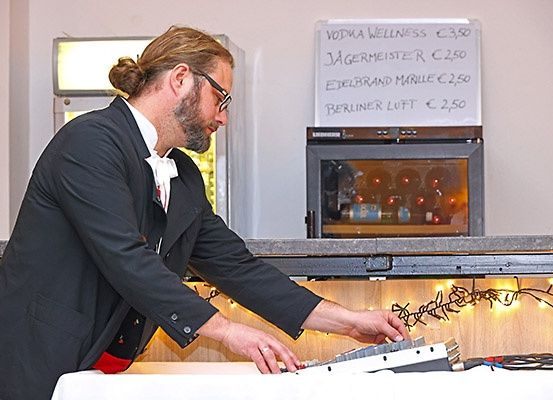 Ein Mann in Anzug und Brille steht an einem Tisch, möglicherweise in einer Bar, und stellt Geräte ein. Hinter ihm befindet sich ein Whiteboard mit Getränkepreisen und ein Kühler mit Flaschen.