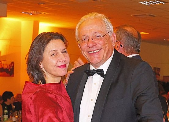 Ein älterer Mann und eine Frau stehen lächelnd für ein Foto bei einer formellen Veranstaltung, mit anderen Gästen und einem dekorierten Raum im Hintergrund.