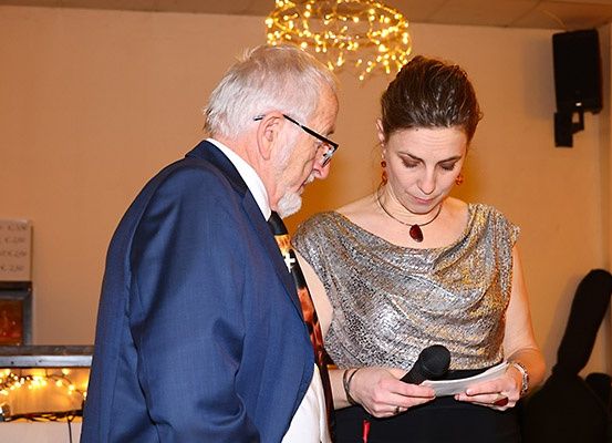 Ein älterer Mann in einem blauen Anzug und mit Brille flüstert etwas einer Frau in einem silbernen Kleid und roten Ohrringen zu, die ein Mikrofon und Papier in der Hand hält, in einem Raum mit warmer Beleuchtung.