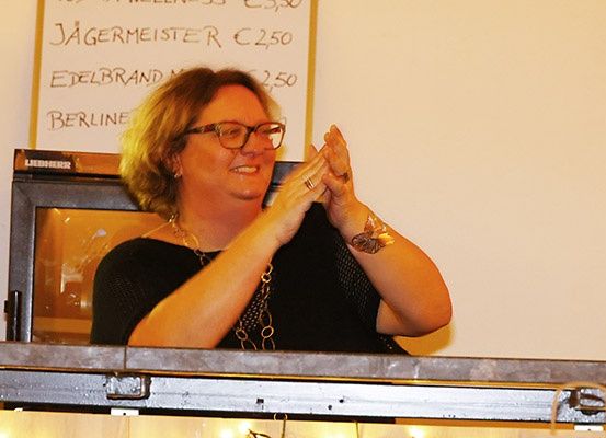 Eine Frau mit Brille und Schmuck sitzt auf der Bühne, klatscht und hat ein Schild mit Preisangaben hinter sich.