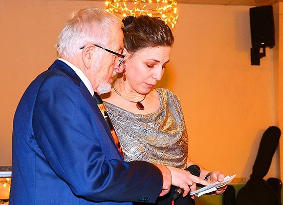 Ein älterer Mann und eine Frau stehen eng beieinander, er schaut sie an und sie hält ein Papier und ein Mikrofon.