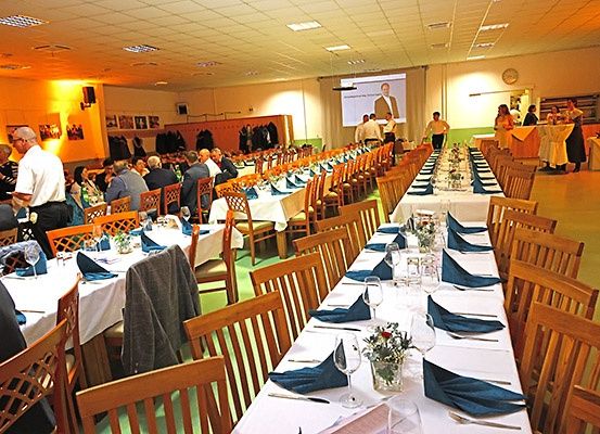 Ein Bankettsaal mit langen Tischen, die für eine große Versammlung gedeckt sind. Jeder Tisch ist mit weißen Tischdecken, blauen Servietten und Weingläsern geschmückt. Die Leute sitzen, unterhalten sich und ein Mann steht auf der Bühne mit einer Projektionsleinwand hinter ihm.