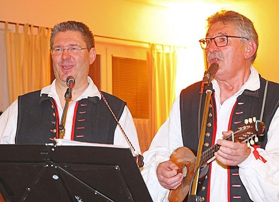 Zwei Männer singen und spielen Gitarre in einem Raum mit einem verhangenen Fenster und einer gelben Wand.