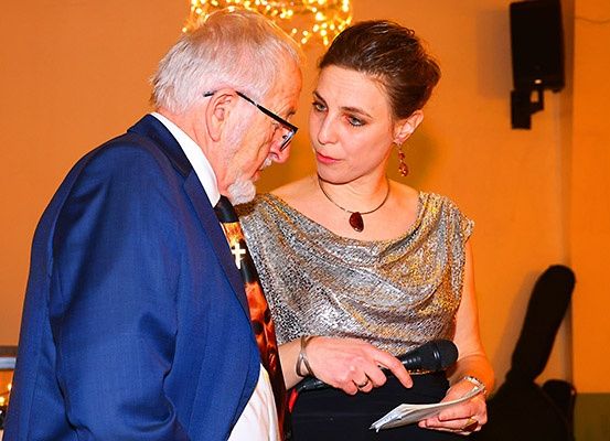 Ein älterer Mann und eine Frau in formeller Kleidung stehen eng beieinander. Die Frau hält ein Mikrofon und Papier und spricht zu dem Mann.