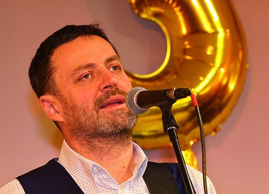 Ein Mann singt in ein Mikrofon vor einem goldenen Luftballon mit der Nummer fünf auf einem rosa Hintergrund.