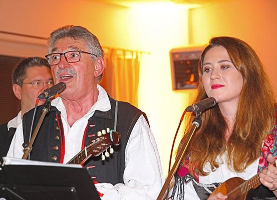 Ein Mann und eine Frau spielen Gitarre und singen in Mikrofone. Sie tragen traditionelle Kleidung. Ein Mann hinter ihnen spielt ebenfalls ein Instrument.