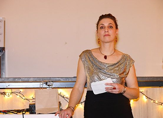 Eine Frau steht an einem Tisch, hält ein Stück Papier und einen Stift in der Hand. Sie ist in einem goldenen Kleid und einem schwarzen Rock gekleidet. Hinter ihr ist eine Bartheke mit einem Weinglas und einer Weinpreisliste zu sehen. Ein Papierbeutel und festliche Lichter sind auf der Theke.