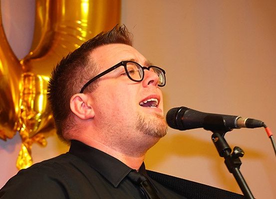 Ein Mann mit Brille und schwarzem Hemd singt in ein Mikrofon, mit einem goldenen Luftballon im Hintergrund.