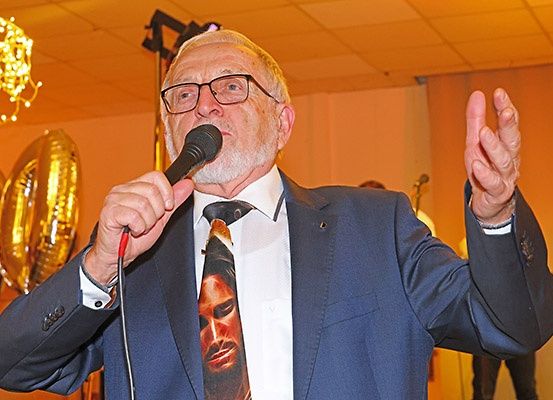 Ein älterer Mann in Anzug und Krawatte spricht in ein Mikrofon mit einer Krawatte, die das Gesicht eines Mannes zeigt. Goldene Ballons und eine Kristallleuchter schmücken den Hintergrund.