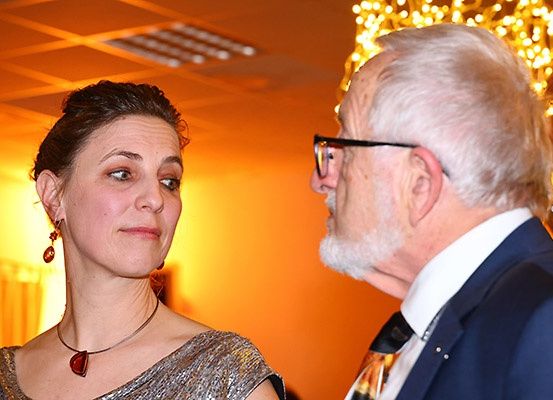 Eine Frau in einem silbernen Kleid und einer Halskette spricht mit einem älteren Mann, der eine Brille, einen Anzug und eine Krawatte trägt.