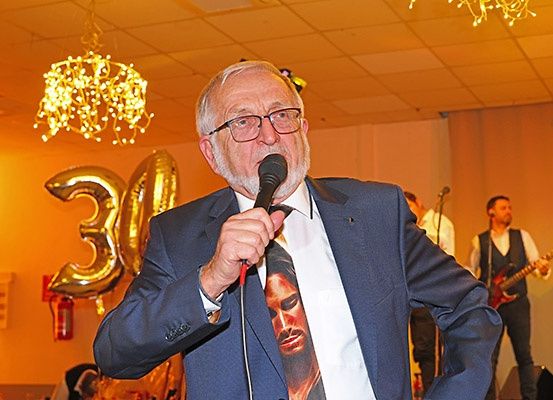 Ein älterer Mann in Anzug und Brille spricht in ein Mikrofon. Hinter ihm befindet sich ein goldener Luftballon mit der Nummer 30. Ein Mann spielt Gitarre im Hintergrund.