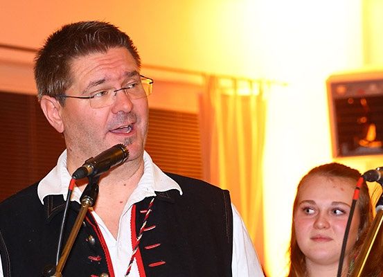 Ein Mann in einer Weste singt in ein Mikrofon, während ein Mädchen zusieht. Sie tragen beide eine Brille.