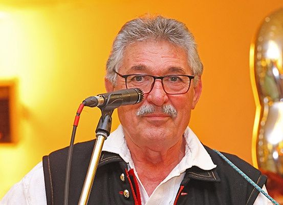 Ein älterer Mann mit Schnurrbart und Brille spricht in ein Mikrofon mit Ständer. Er trägt einen schwarzen Weste und ein weißes Hemd. Hinter ihm ist eine gelbe Wand und ein Bilderrahmen.