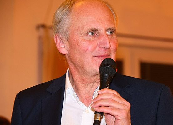 Ein älterer Mann in einem Anzug hält ein Mikrofon und spricht bei einer Veranstaltung, mit einem verschwommenen Publikum im Hintergrund.