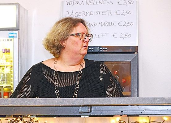 Eine Frau mit kurzen blonden Haaren und Brille steht an einer Theke. Hinter ihr befindet sich ein Speiseplan und ein Kühlschrank.