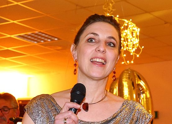 Eine Frau in einem goldenen Kleid spricht in ein Mikrofon bei einer Veranstaltung. Hinter ihr ist ein großer goldener Ballon zu sehen, und zwei Männer, einer mit Brille, beobachten.