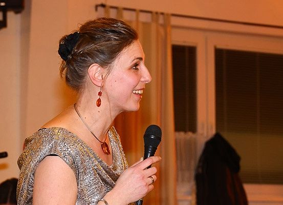 Eine Frau mit braunen Haaren, die ein silbernes Kleid und rote Ohrringe trägt, hält ein Mikrofon und scheint bei einer Veranstaltung zu sprechen. Im Hintergrund befinden sich Fenster mit Vorhängen und eine Jacke, die an einer Tür hängt.