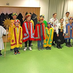 Eine Gruppe von Kindern in bunten Kostümen, möglicherweise für ein Theaterstück, stehen in einem Raum mit grünem Boden.