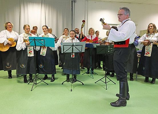 Eine Gruppe von Menschen in traditioneller Kleidung steht in einem Raum und spielt Musikinstrumente. Ein Mann vor ihnen spielt Gitarre.