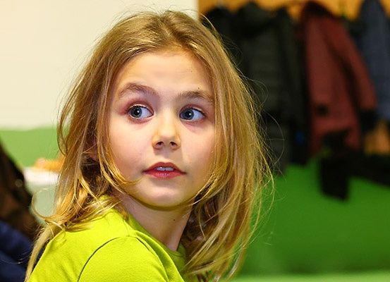Ein junges Mädchen mit blonden Haaren und einem grünen T-Shirt sieht überrascht aus, möglicherweise in einem Klassenzimmer mit unscharfem Hintergrund.