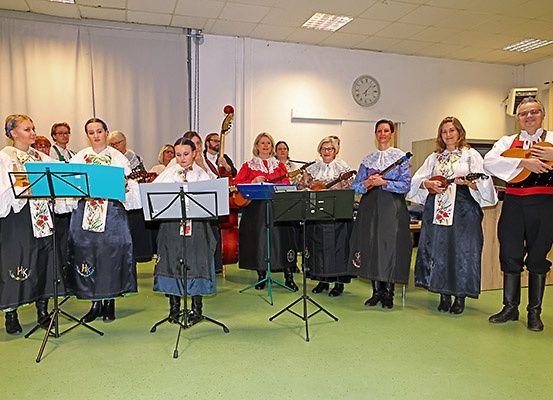 Eine Gruppe von Musikern in traditioneller Kleidung, die in einem Raum mit Notenständern stehen, spielen verschiedene Instrumente wie Gitarren und Cello. Eine Uhr und Vorhänge sind im Hintergrund zu sehen.
