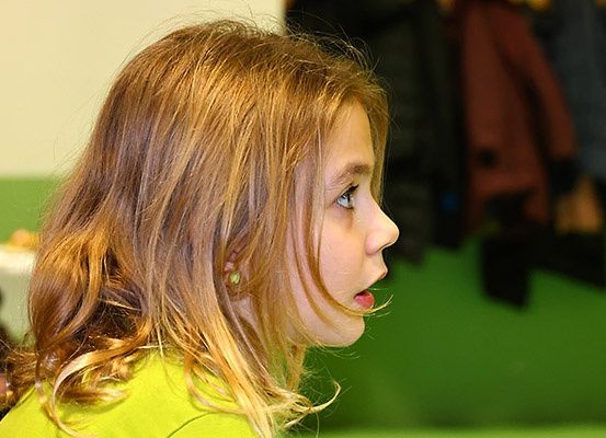 Ein junges Mädchen mit blonden Haaren und grünen Ohrringen schaut mit offenem Mund zur Seite. Hinter ihr befindet sich eine grüne Wand und Kleidung, die an der Wand hängt.