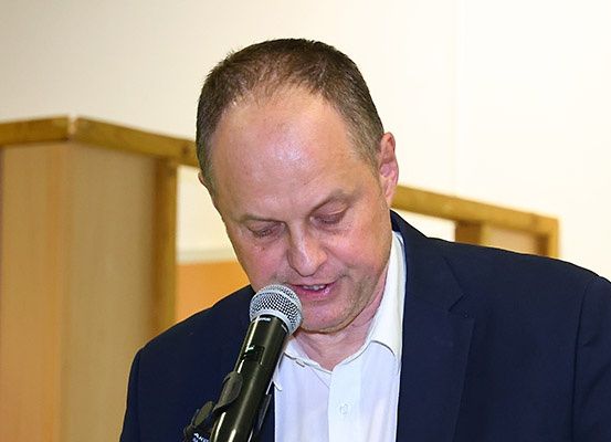 Ein Mann in einem dunkelblauen Anzug spricht in ein Mikrofon und schaut nach oben, als wäre er in Gedanken. Hinter ihm ist eine Holzwand.