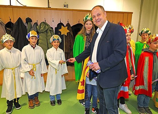 Ein Mann überreicht einer Gruppe von Kindern in Kostümen ein Geschenk. Die Kinder lächeln, und der Mann trägt einen Anzug. Hinter ihnen hängen Jacken an einer Holzwand.