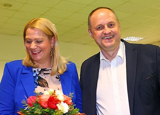 Ein Mann und eine Frau lächeln und stehen vielleicht für ein Foto eng zusammen. Die Frau hält einen Blumenstrauß. Sie sind beide formell gekleidet.