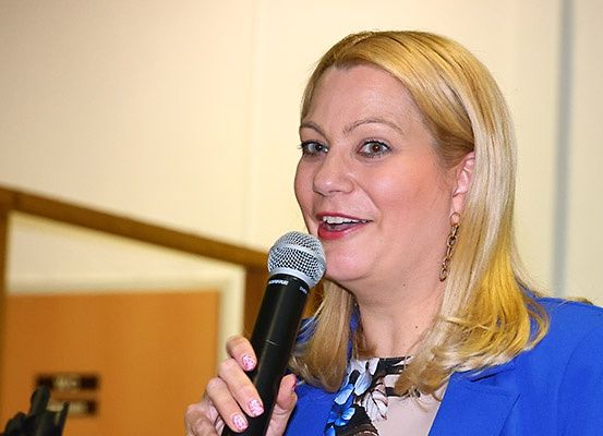 Eine Frau mit blondem Haar lächelt, während sie ein Mikrofon in einem Konferenzraum hält. Sie trägt einen blauen Blazer und ein Blumenoberteil.