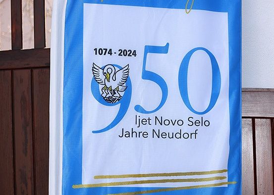 Blaue Fahne mit '950', Vogellogo, Jahren '1074-2024' und Text 'Ijet Novo Selo Jahre Neudorf'.